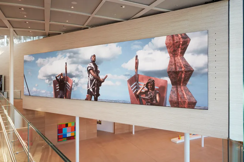Two-story gallery interior with a large horizontal digital screen mounted on a timber-clad wall above the lower level