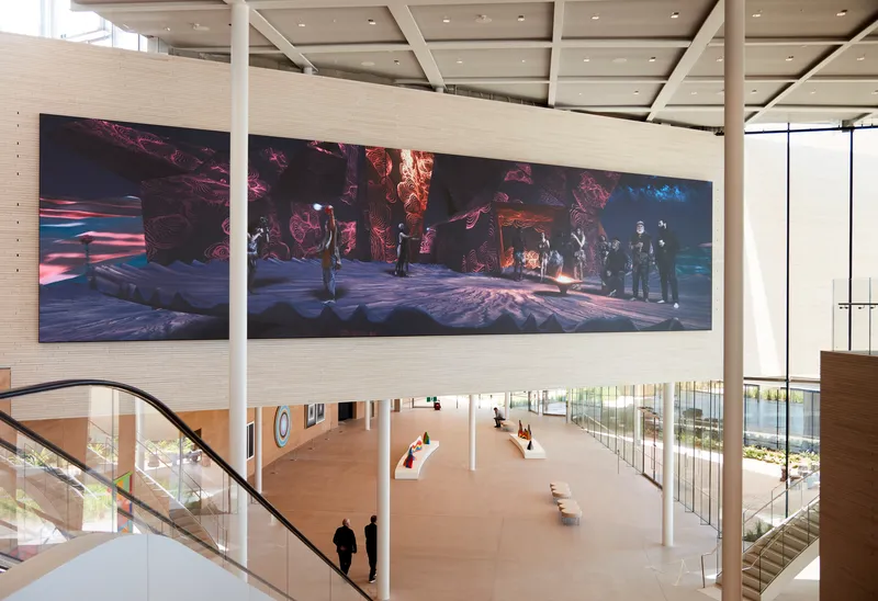 Double-height gallery space with a person descending a curved staircase beneath a wide horizontal video projection