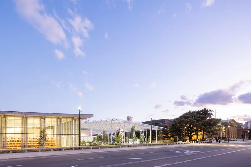 Street view of the glass-walled entrance pavilion and lattice roof structure at dusk with palm trees visible beyond