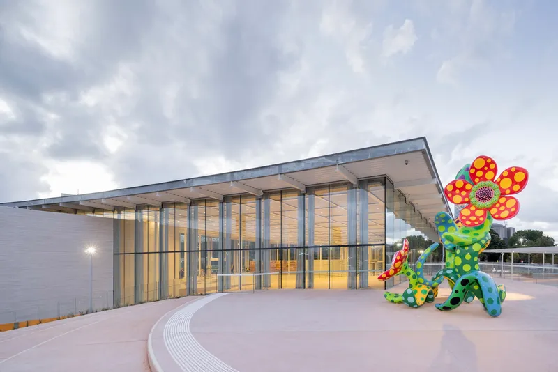 Glazed entrance facade with a wide overhanging roof and a colorful sculptural installation on the plaza at twilight