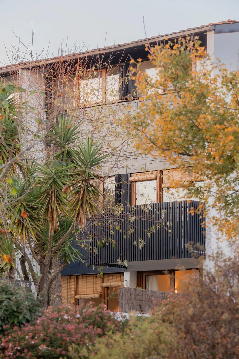 Three-story facade with corrugated metal balcony screen framed by autumn foliage and palm fronds