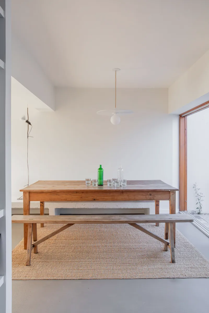Timber dining table and benches on sisal rug beneath brass pendant light in white-walled room