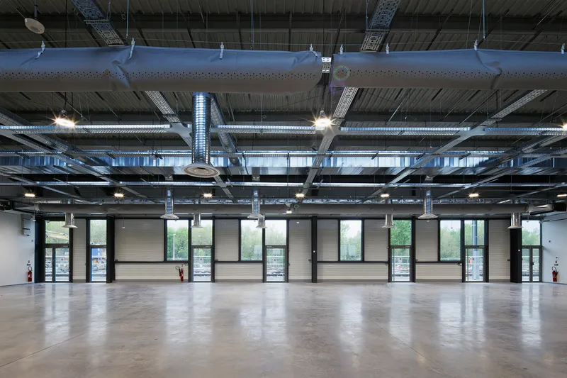 Open-plan interior space with exposed ceiling ducts, pendant lights, and a glazed facade overlooking greenery