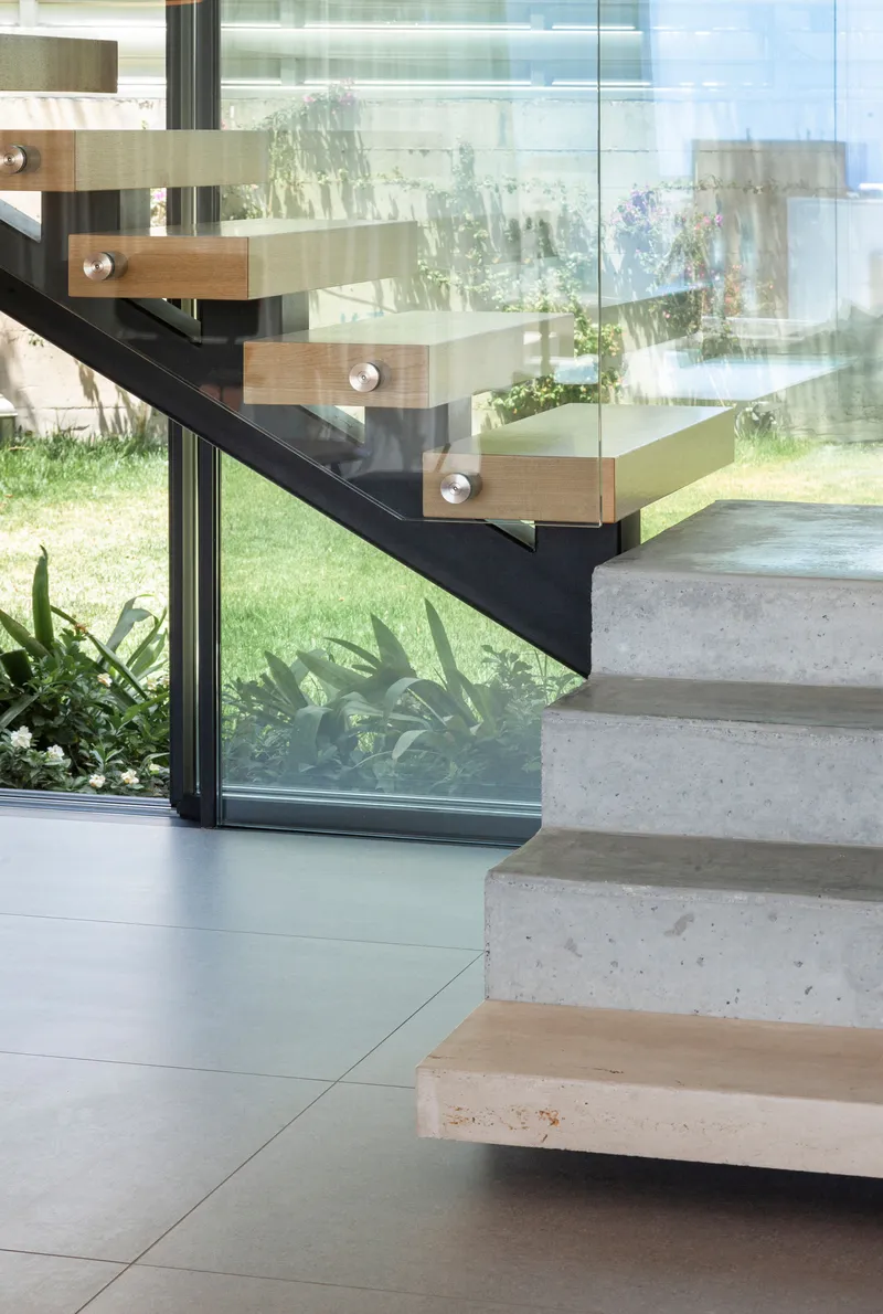 Concrete treads with timber nosings and black steel stringer beside floor-to-ceiling glass wall with garden beyond