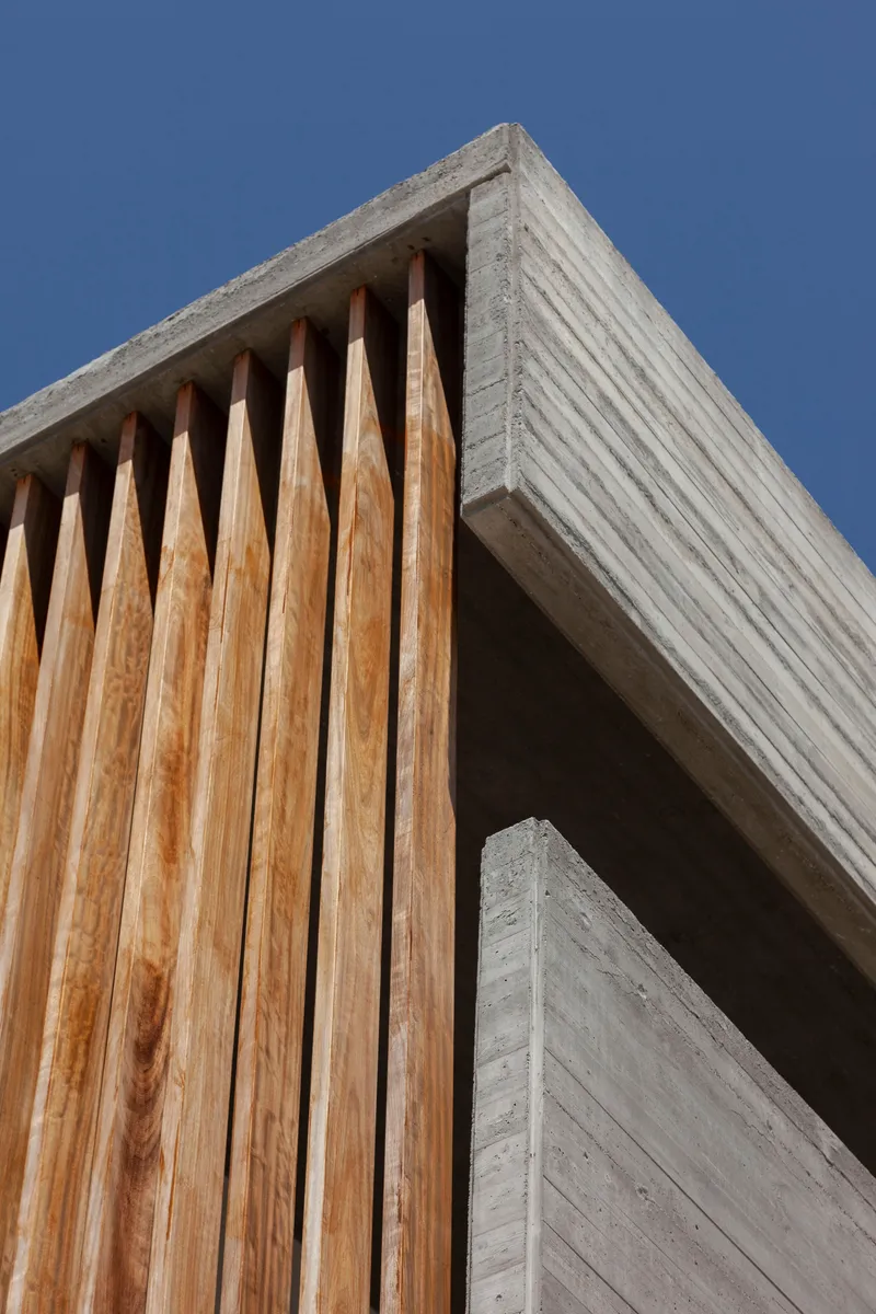 Close-up of board-formed concrete beam corner meeting vertical timber slats under bright daylight