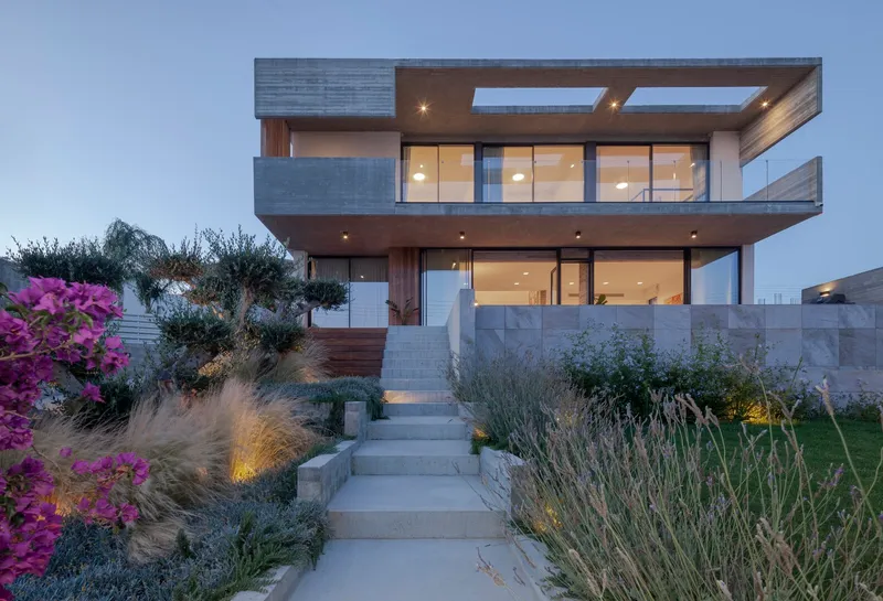 Three-level cantilevered residence with lit interiors at dusk accessed by concrete stairs through planted gardens