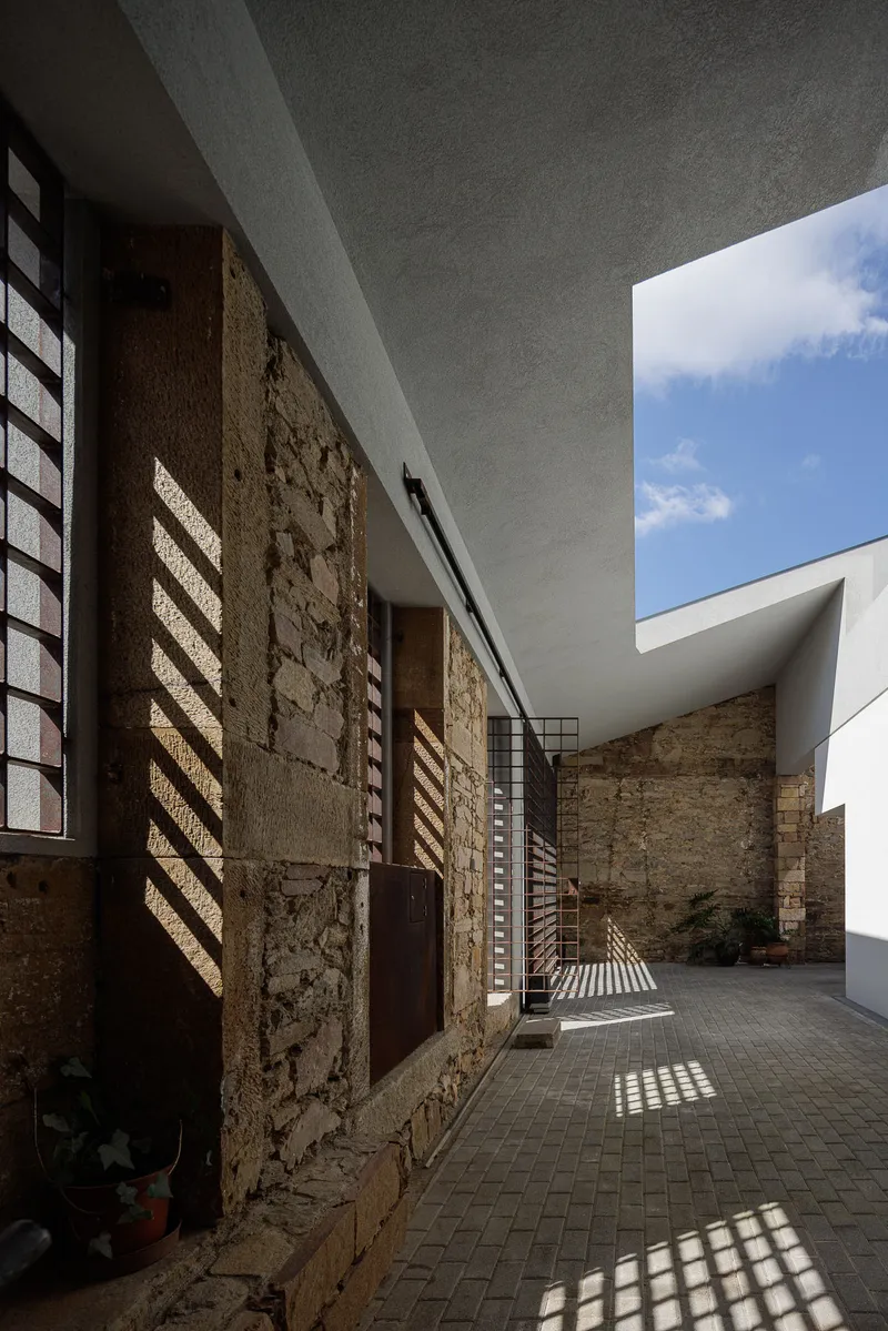 Narrow courtyard with weathered stone wall and gridded metal screen creating striped sunlight patterns
