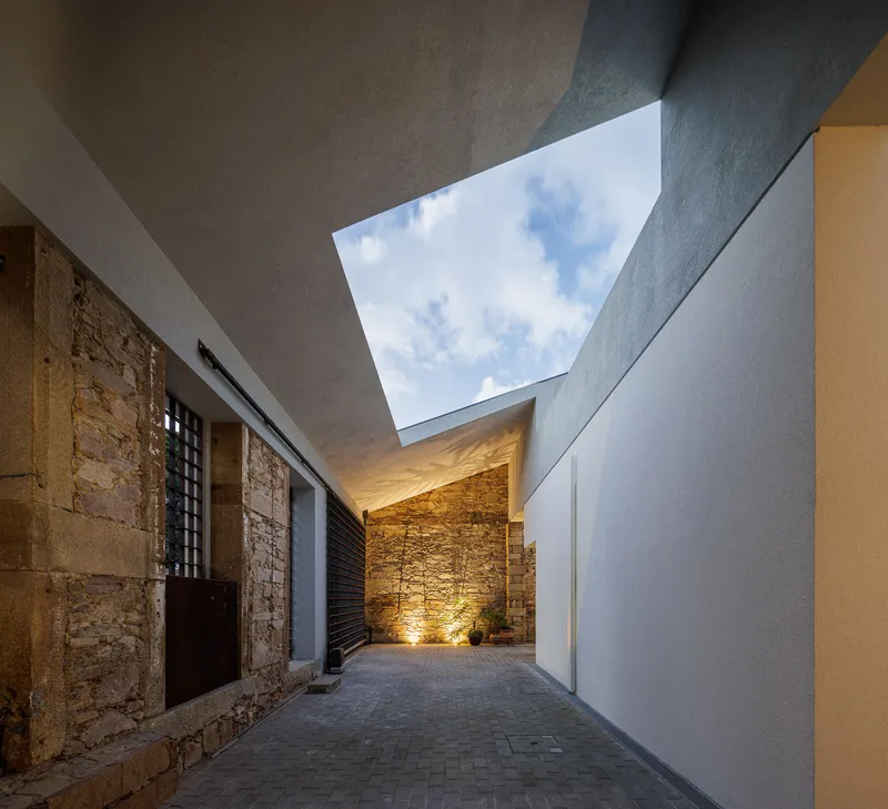Courtyard passageway with rectangular skylight opening framing blue sky and uplighting on stone wall at dusk