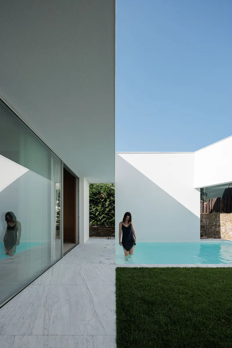White courtyard with rectangular pool and two figures wading in clear water beside glass doors