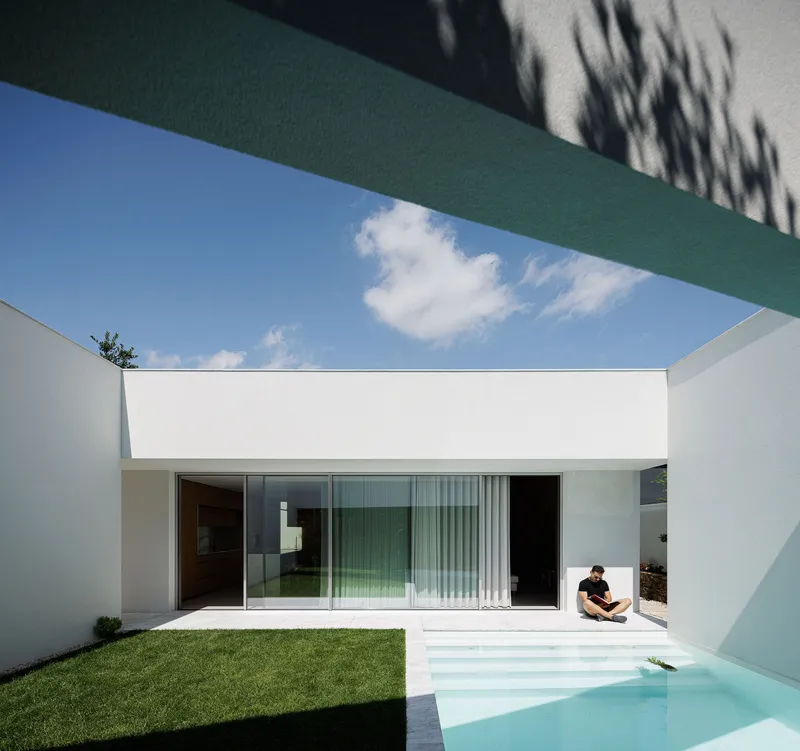 Courtyard pool framed by green-painted soffit with person seated at water's edge beneath blue sky