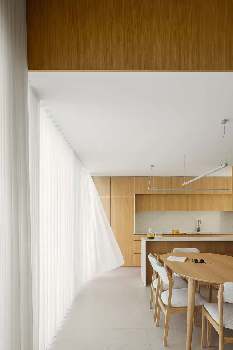Dining area beneath a timber soffit with sheer curtains filtering daylight along the exterior wall