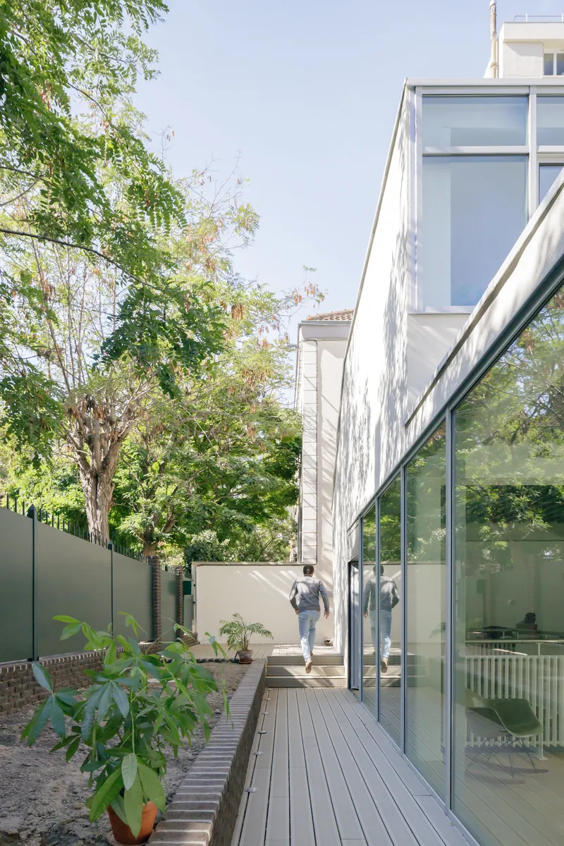 Exterior timber deck walkway alongside glazed facade with figure walking past potted plants and tree canopy above