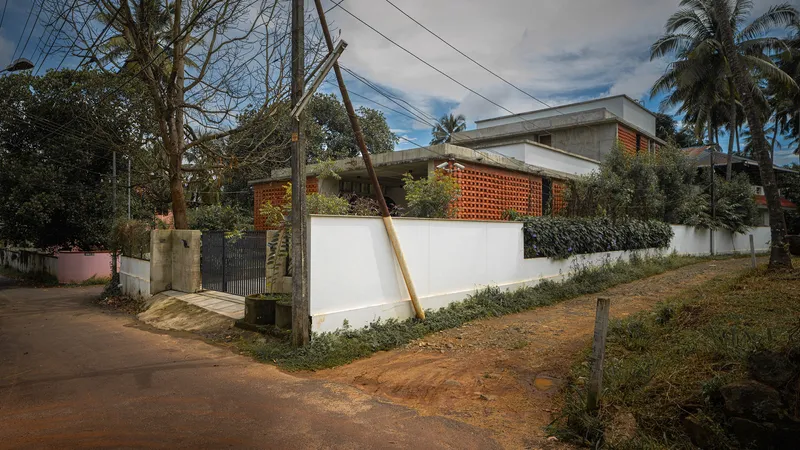Street view showing the white compound wall, concrete roof, and perforated brick volumes among palm trees and utility poles