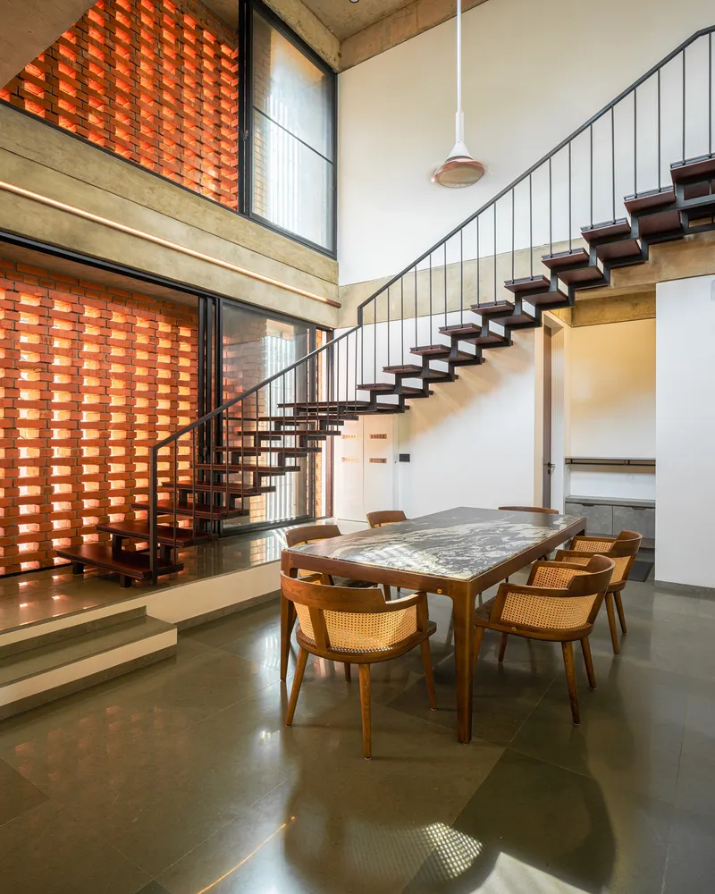 Dining area with floating timber staircase, suspended pendant light and double-height brick screen wall filtering sunlight