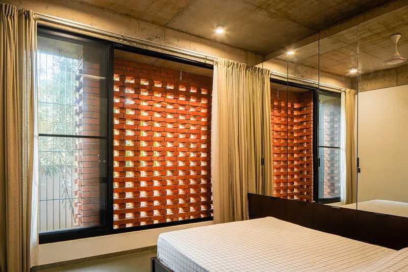 Bedroom with perforated brick window screen, floor-length curtains and exposed concrete ceiling at dusk