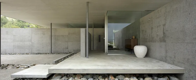 Elevated concrete entry platform floating above river stones with glass walls revealing interior spaces