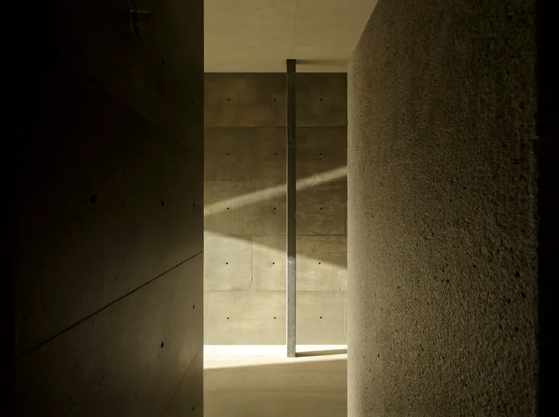 Narrow hallway in board-formed concrete with vertical slot window casting sunlight on textured stone wall