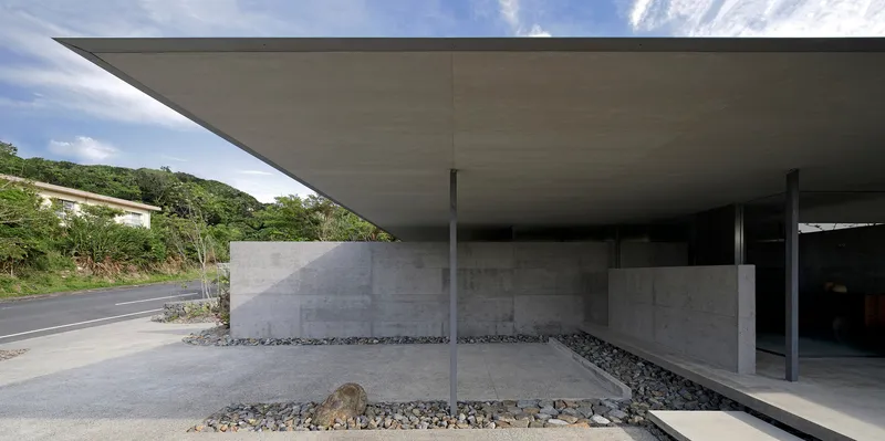 Covered entrance with thin steel columns and concrete walls set in a gravel courtyard beside a hillside road