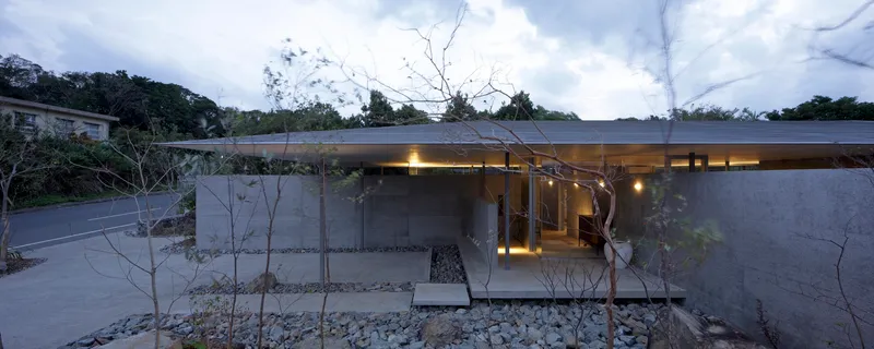 Glass-walled entrance pavilion illuminated at dusk with gravel landscaping and bare winter trees