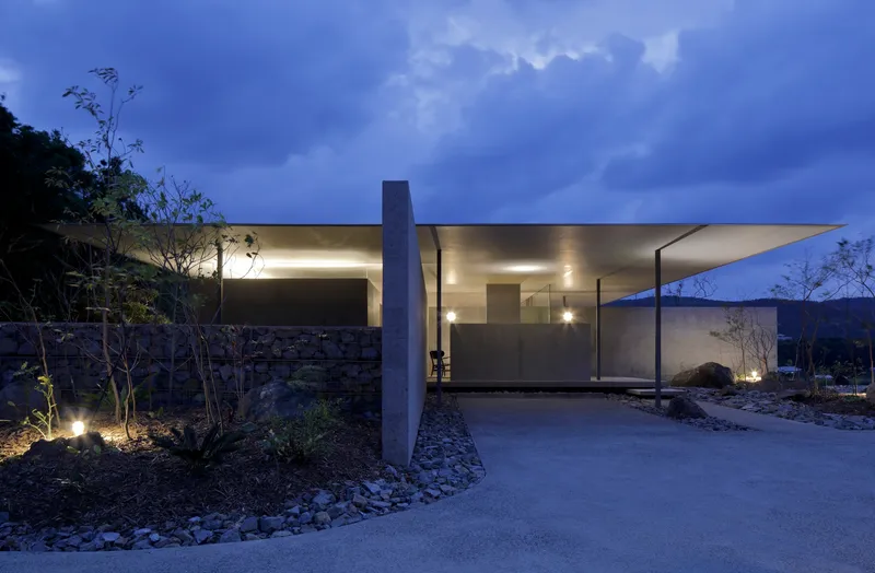 Cantilevered roof plane over glazed and concrete volumes at twilight with uplighting on surrounding vegetation