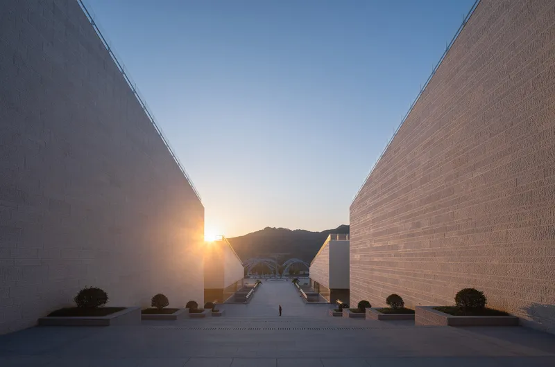 Paved courtyard framed by two angled brick-clad walls converging toward a distant hillside at sunset
