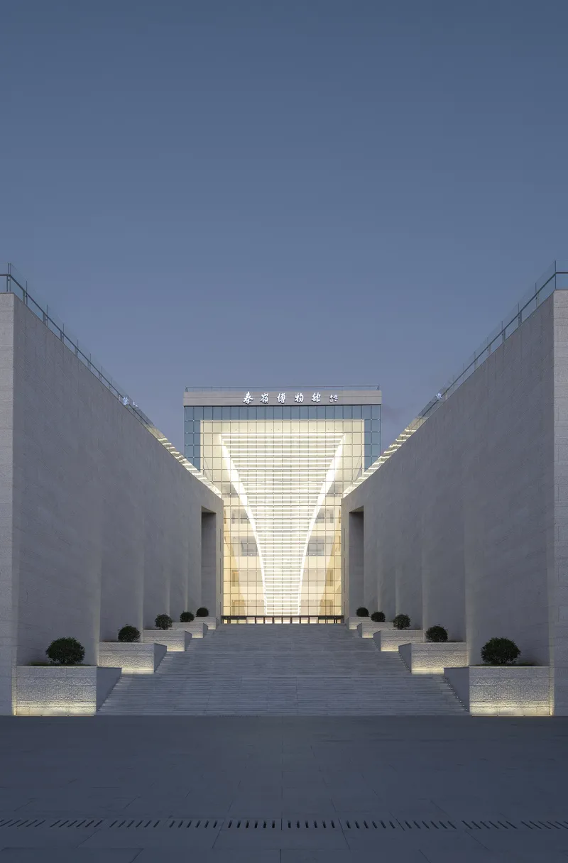 Stone-paved plaza flanked by angled walls leading to the illuminated glass entrance at dusk