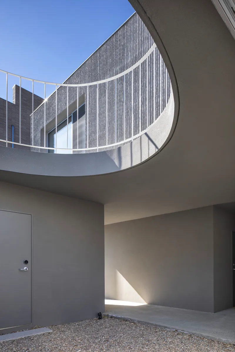 Curved balcony opening with white metal railing overlooking a gravel courtyard with vertical concrete cladding beyond