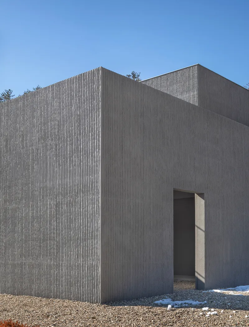 Exterior corner showing vertical concrete cladding with a narrow doorway and gravel ground under clear skies