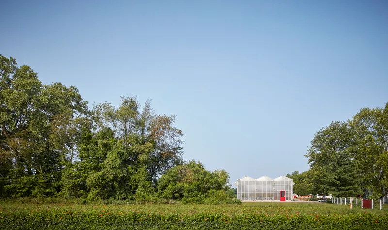 Greenhouse pavilion with translucent polycarbonate walls and red doors viewed across a green field beneath mature trees