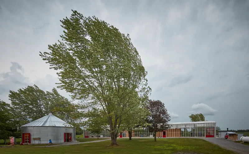 Campus of converted silo and greenhouse buildings beneath windswept trees and stormy afternoon clouds