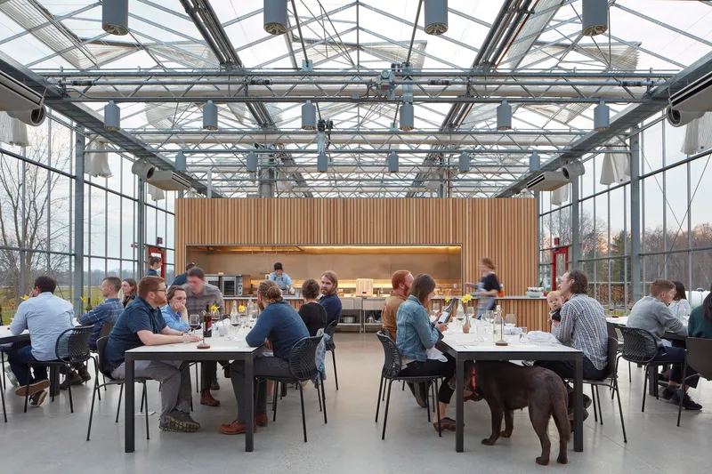 Interior dining area beneath exposed steel truss roof with vertical wood slat service core and seated guests