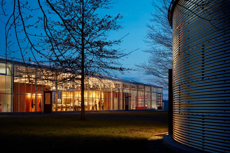 Glass-walled pavilion glowing from interior lights at dusk with bare trees and a cylindrical metal form nearby