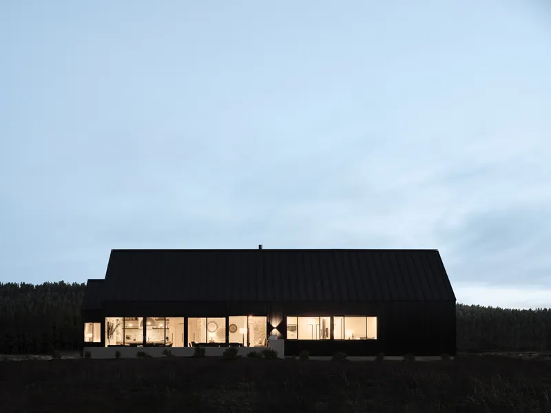 Glazed facade illuminated at night revealing the timber-lined interior and forest backdrop