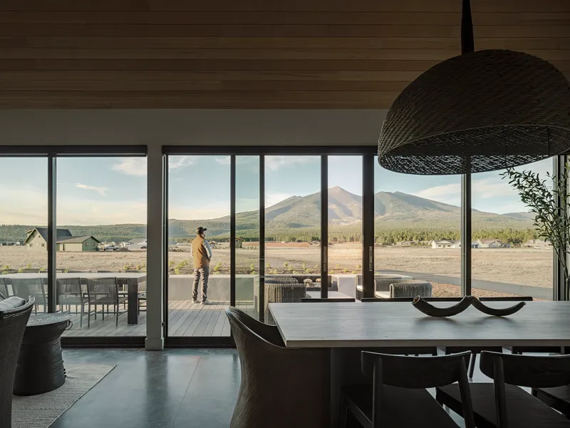 Dining area with full-height glazing and a figure standing on the terrace framed by distant peaks