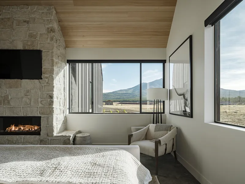 Bedroom with corner windows framing mountain views and stone fireplace with timber ceiling
