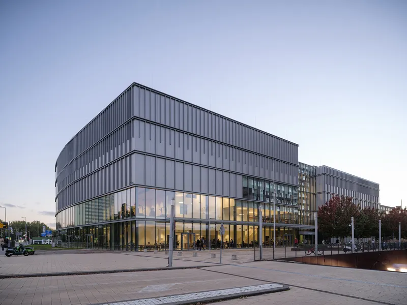 Vertically ribbed metal facade with glazed ground floor facing a paved public plaza at dusk
