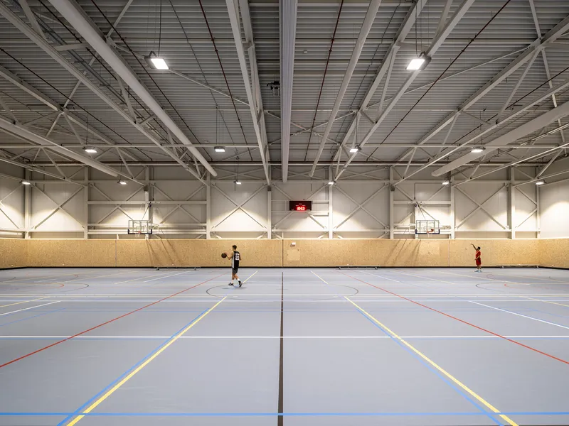 Indoor sports hall with exposed white steel truss structure and plywood wainscoting with two players on court