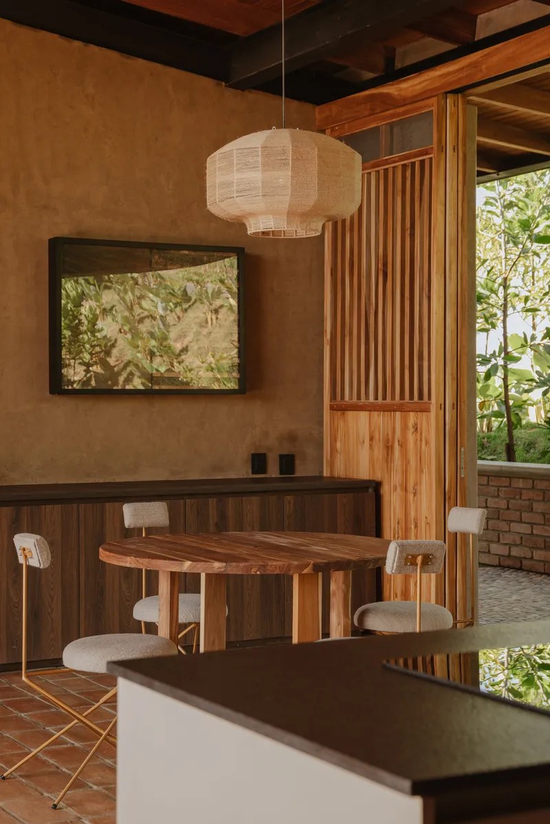 Dining area with timber table, slatted screen partition, and paper pendant light beside rammed earth wall