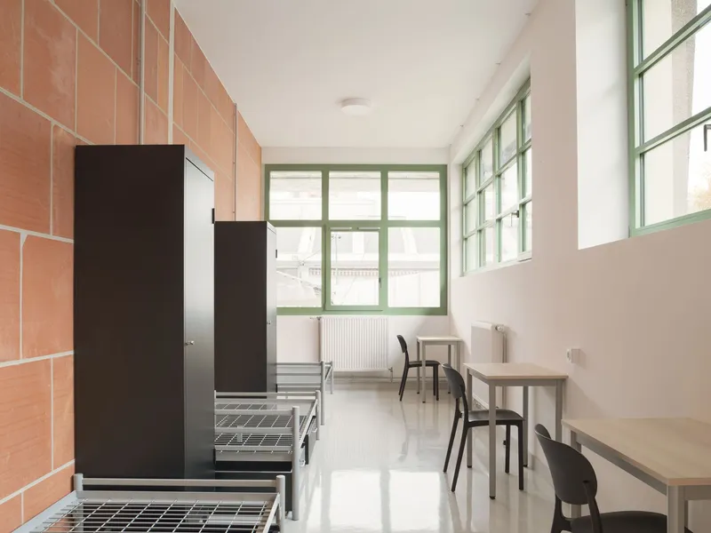 Institutional room with terracotta tile wall, metal bed frames, and green-framed windows overlooking adjacent buildings