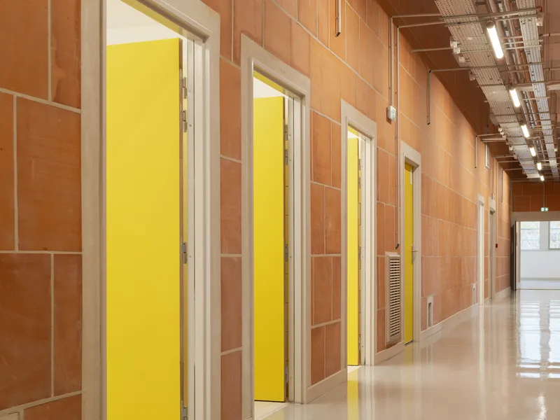 Corridor lined with yellow painted doors set into terracotta tile wall with exposed ceiling services