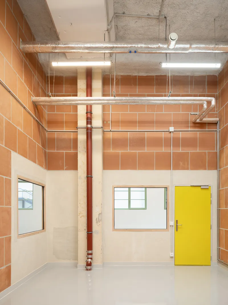 Interior room showing exposed concrete ceiling, terracotta tile walls, red drainage pipe, and yellow door