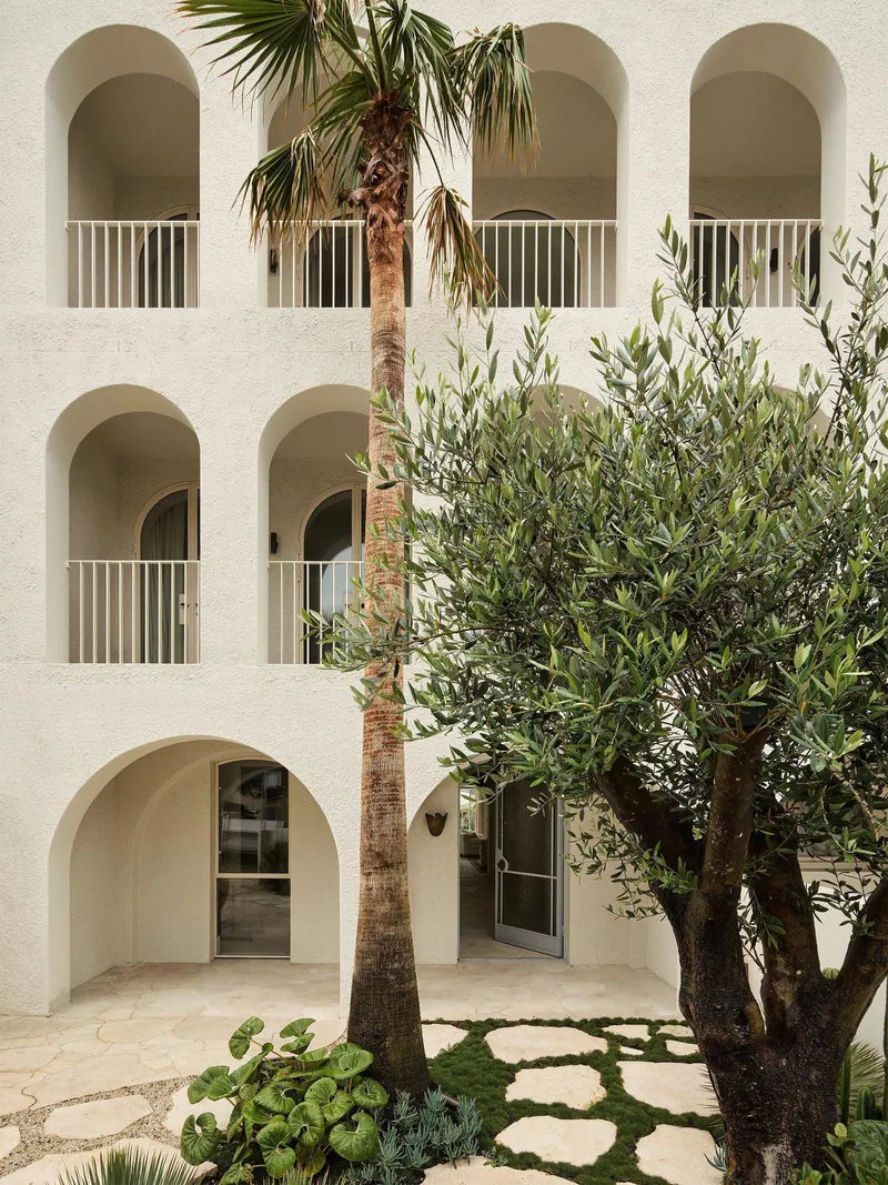 Courtyard view of arched loggias with white railings and planted bed with palm and olive trees