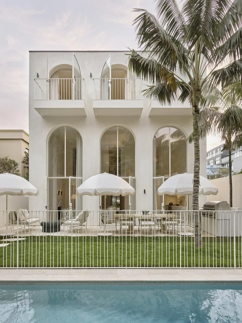 White stucco facade with arched openings and upper balconies beside a pool and palm trees