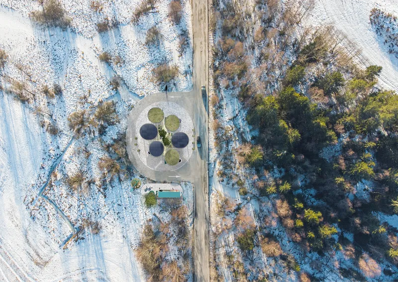 Aerial view of a circular pavilion with four lobed openings surrounded by snow-dusted terrain and evergreen trees