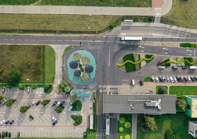 Overhead view of a circular pavilion with turquoise surfacing at a roadside intersection bordered by parking lots