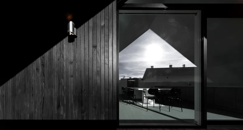 Triangular window reflecting interior dining area at night with exterior sconce illuminating charred timber cladding
