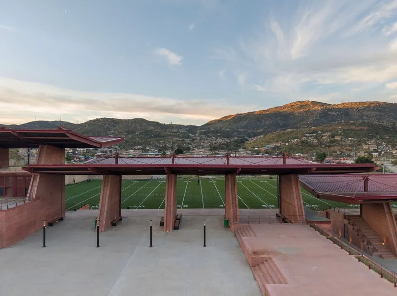 Covered concrete plaza overlooking the playing field with angular roof structures and terraced seating at dusk