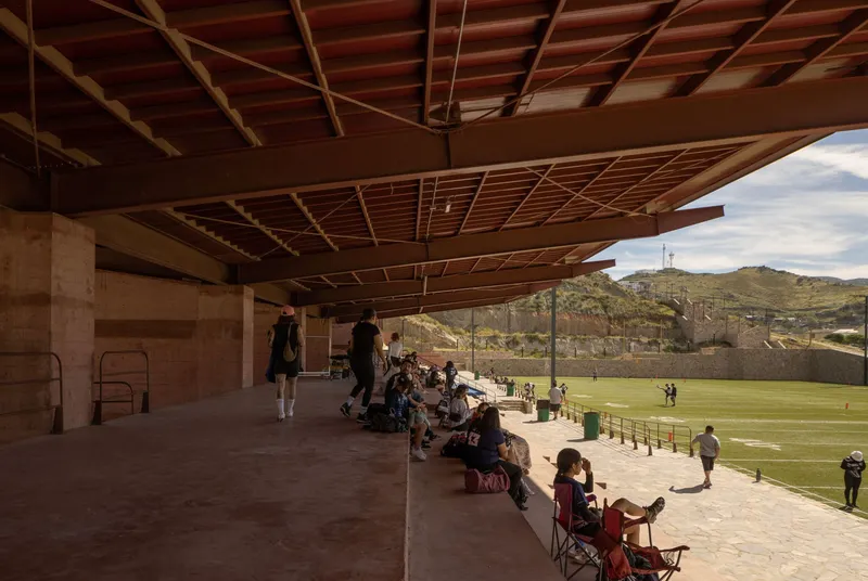 Open-air concourse with exposed timber joists and spectators seated on benches overlooking the field