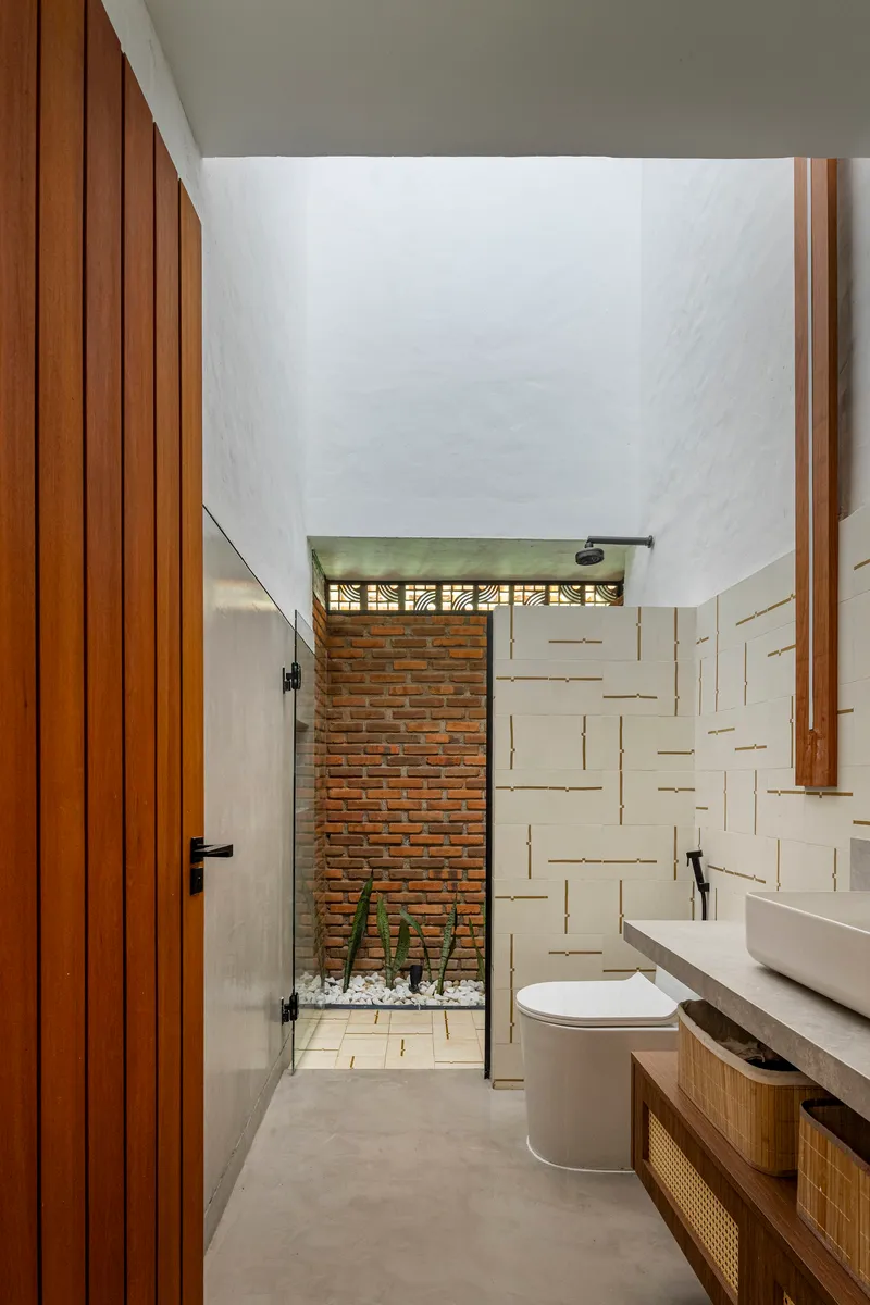 Narrow bathroom with tall ceiling and glass door opening to a brick-walled courtyard with planted gravel bed