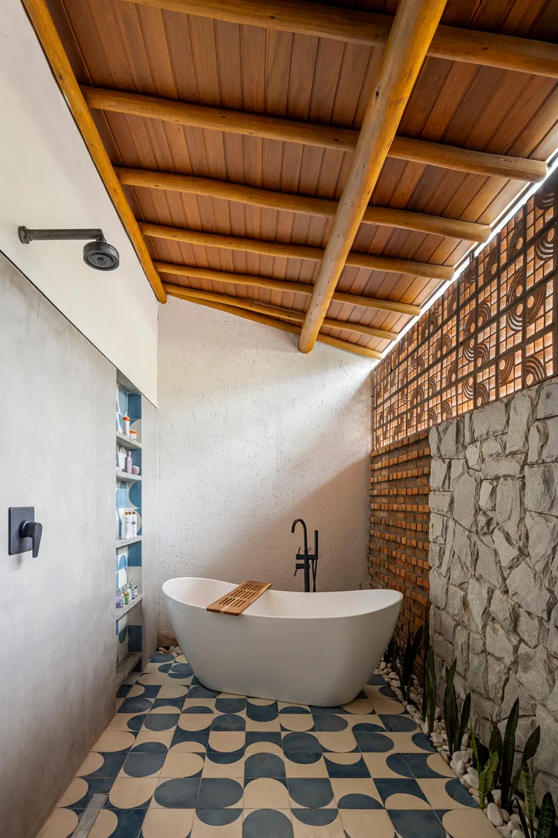 Freestanding bathtub on patterned tile floor beneath exposed timber ceiling beams and latticed brick screen